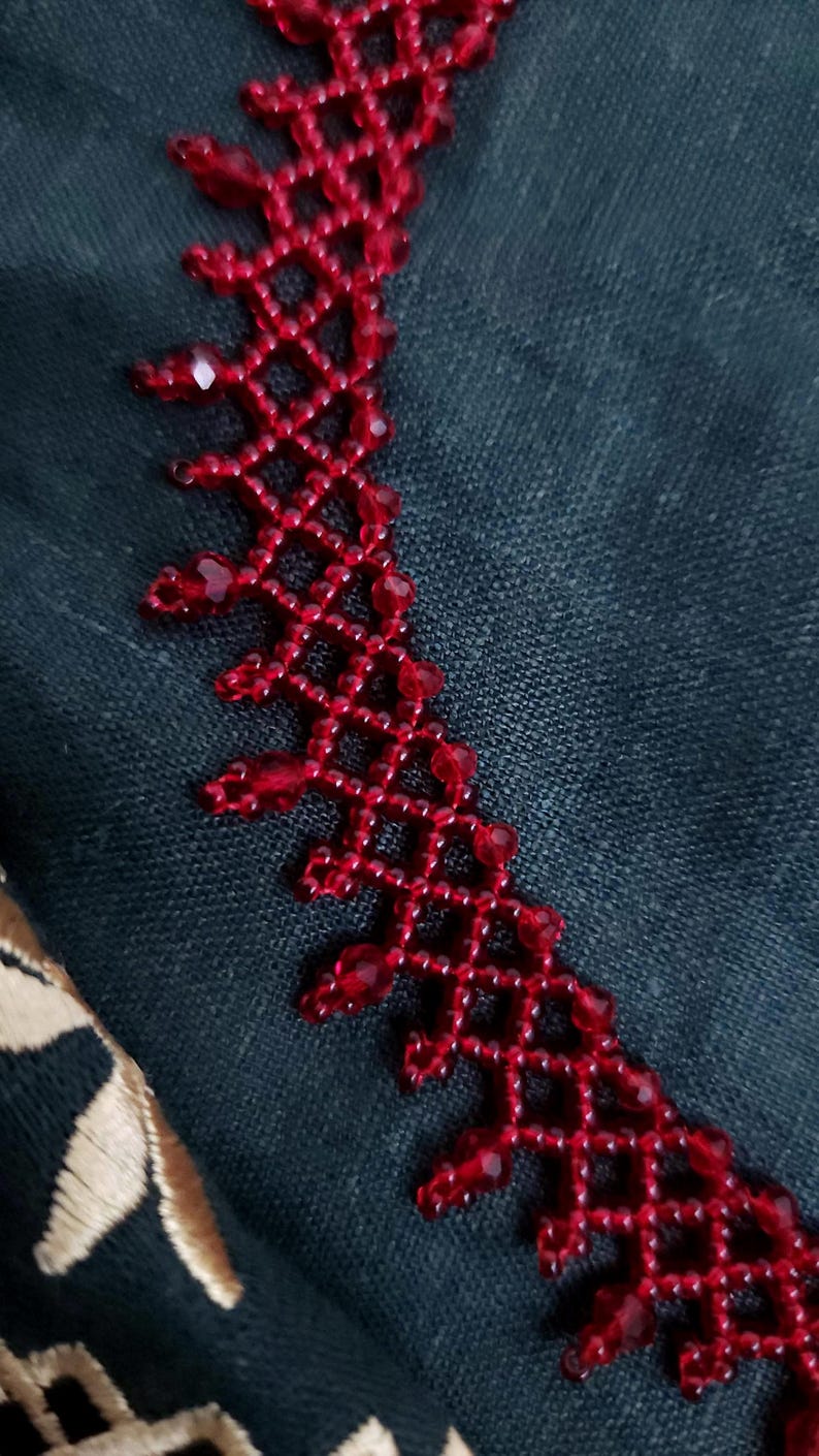 May include: Close-up of a red beaded necklace with a geometric design. The necklace is made of small, faceted beads and is set against a dark teal fabric background. The intricate beadwork creates a delicate, lacy effect.