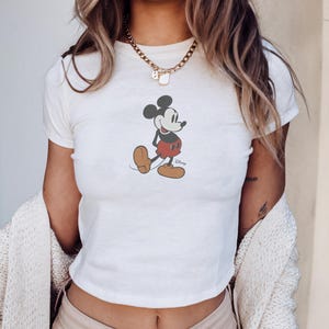 May include: White crop top featuring a classic Mickey Mouse graphic. The cartoon character is depicted in his iconic pose, wearing red shorts, brown shoes, and white gloves. The Disney logo is visible below the character.