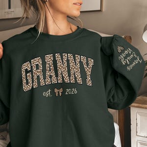 May include: Forest green sweatshirt with the word "GRANNY" in a leopard print design. The sweatshirt also features the text "est. 2026" and the names "James" and "Emily Noah" on the sleeve. The sweatshirt is a comfortable, casual style.