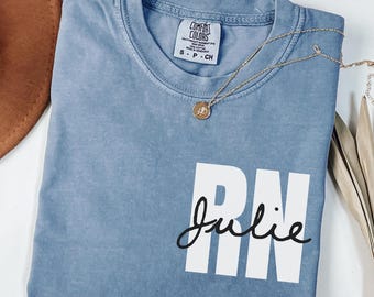 May include: Blue jean colored t-shirt with the text "RN Julie" in white and black. The shirt has a crew neck and short sleeves. A gold necklace with a pendant is draped across the shirt.