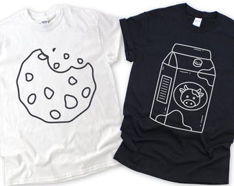 May include: Two t-shirts: one white, one black. The white shirt features a black outline of a bitten cookie. The black shirt displays a white outline of a milk carton with a cow graphic. Both shirts are short-sleeved.