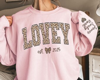 May include: Light pink sweatshirt with the word "LOVEY" in large, leopard print letters across the chest. Below the word "LOVEY" is the text "est. 2025" with a bow graphic. The right sleeve has the names "James, Emily, Noah" and a heart.