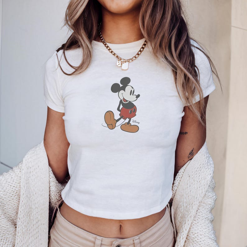May include: White crop top featuring a classic Mickey Mouse graphic. The cartoon character is depicted in his iconic pose, wearing red shorts, brown shoes, and white gloves. The Disney logo is visible below the character.
