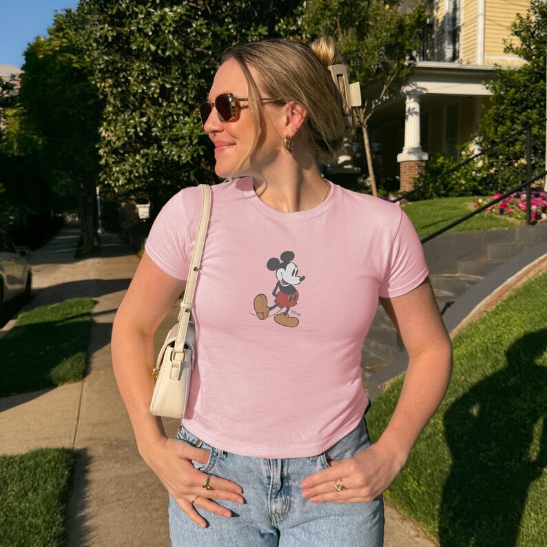 May include: A light pink t-shirt featuring a graphic of Mickey Mouse in a walking pose. The shirt has short sleeves and a crew neck. The person is wearing blue jeans, sunglasses, and a white purse.