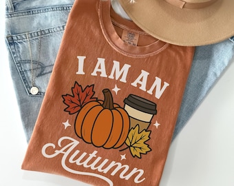 May include: An orange t-shirt with the text "I AM AN Autumn" and a graphic of a pumpkin, coffee cup, and leaves. The shirt is laid on top of blue jeans, a tan hat, and a gold necklace.