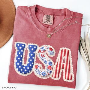 May include: Crimson-colored t-shirt with the word "USA" appliqued on the front. The letters are decorated with patriotic patterns: blue stars, floral print, and red and white stripes. A silver necklace with a small pendant is draped across the shirt.