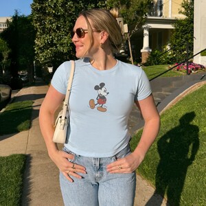 May include: Light blue t-shirt featuring a classic Mickey Mouse graphic. The shirt has short sleeves and a crew neck. The person is wearing light blue jeans and sunglasses. A white purse is hanging from the shoulder.