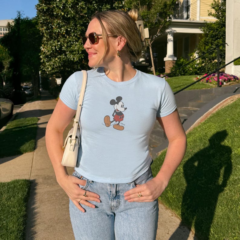 May include: Light blue t-shirt featuring a classic Mickey Mouse graphic. The shirt has short sleeves and a crew neck. The person is wearing light blue jeans and sunglasses. A white purse is hanging from the shoulder.