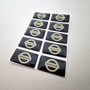 Op de afbeelding: Een vel met zwarte en witte Nissan autologo stickers. De stickers zijn glanzend en hebben een reliëf 3D-effect.