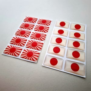 May include: Two sheets of stickers with a red circle and white rays design. One sheet has 12 stickers with a red circle and white rays design. The other sheet has 10 stickers with a red circle on a white background.