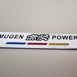 May include: A rectangular, brushed metal emblem with the text "MUGEN POWER" in black. The emblem also features Japanese characters and three horizontal stripes in blue, red, and gold.