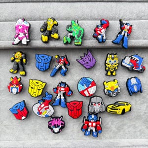 May include: A collection of twenty-four colourful rubber shoe charms featuring various robot characters, vehicle forms, and faction insignias in red, blue, yellow, pink, green, purple, and black, arranged in rows on a grey textured surface.