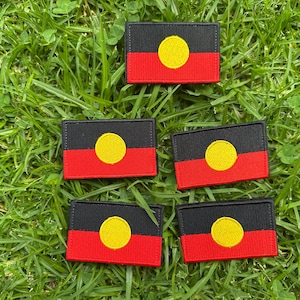 May include: Five rectangular patches displaying the Aboriginal flag design. Each patch features a black upper section, a red lower section, and a yellow circle in the centre. The patches are set on green grass.