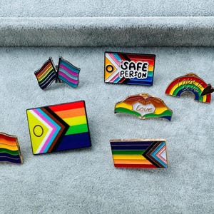 May include: A set of ten enamel pins featuring various LGBTQ+ pride flags and symbols. The pins include a rainbow flag, a transgender flag, a bisexual flag, a progress pride flag, a safe person flag, a rainbow heart, a rainbow with the text "This is love", a rainbow heart with the text "Love", and a rainbow flag with a chevron design.