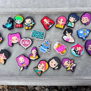 May include: A selection of colourful shoe charms with cartoon characters and designs. The charms feature anime-style figures, hearts, and the text "K-POP DEMON HUNTERS". The charms are made of a rubbery material.