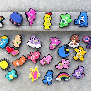 May include: A collection of colourful rubber shoe charms featuring various Care Bears characters, rainbows, hearts, a sun, a moon, and clouds. Each charm is detailed with vibrant colours and distinct shapes, designed to be attached to footwear accessories.