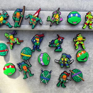 May include: A collection of colourful rubber shoe charms featuring various Teenage Mutant Ninja Turtles characters in action poses, along with individual turtle shells and head portraits in green, red, blue, orange, and purple colour schemes.