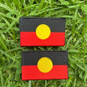 May include: Two rectangular patches displaying the Aboriginal Australian flag. Each patch has a black upper section, a red lower section, and a yellow circle in the centre. The patches are placed on a bed of green grass.