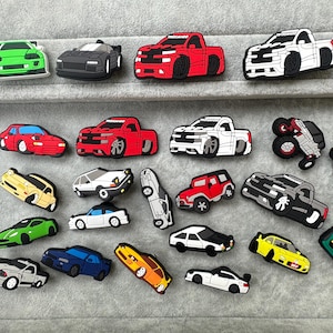 May include: A selection of car-shaped accessories in various colours, including green, black, red, white, and yellow. The collection features different car models, such as trucks, sports cars, and saloons, in a variety of designs.