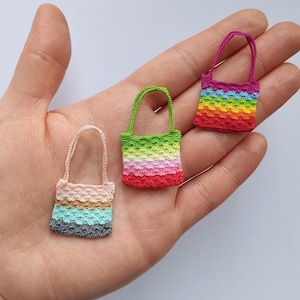 May include: Three miniature crocheted tote bags in a hand. The bags feature colorful striped patterns in pink, green, red, blue, and rainbow hues. Each bag has a matching handle. The bags are small, decorative, and likely handmade.