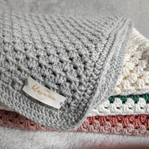 Op de afbeelding: Een stapel handgemaakte gehaakte dekens in verschillende kleuren. De bovenste deken is grijs, gevolgd door crème en een roze onderaan. Een klein label met het woord "blasmeria" is bevestigd aan de grijze deken.