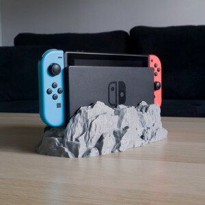 May include: A 3D printed grey rock-shaped stand for a Nintendo Switch console and two controllers. The console is black and the controllers are blue and red.