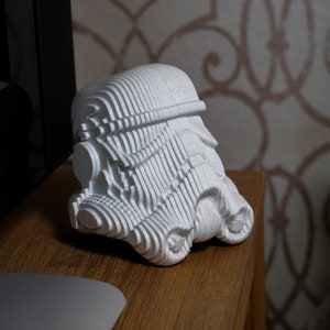 Peut inclure: Modèle d'un casque de Stormtrooper de Star Wars imprimé en 3D, blanc et en couches. Le casque est posé sur une surface en bois.