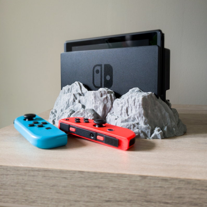 May include: A grey, 3D printed mountain range stand for a Nintendo Switch console. The console is black and has the Nintendo Switch logo on the front. Two controllers are on the table, one red and one blue.
