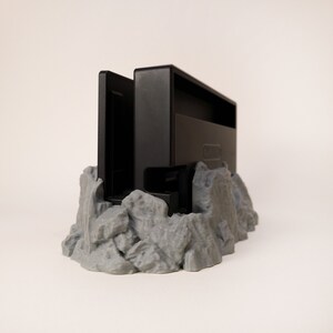 May include: A black Nintendo Switch game cartridge holder with a grey, rocky base. The holder is designed to look like a mountain range.