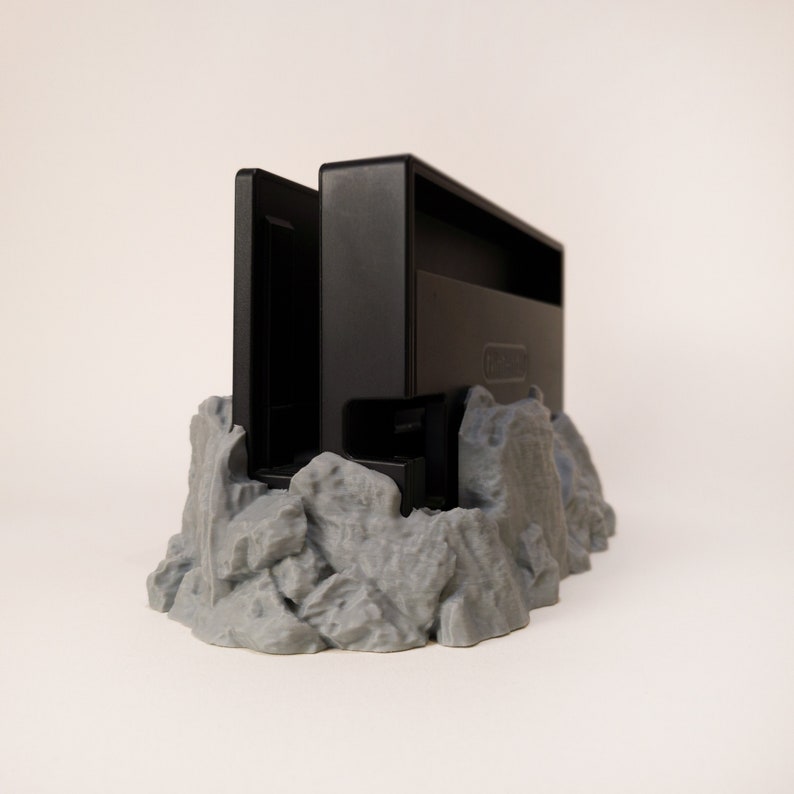 May include: A black Nintendo Switch game cartridge holder with a grey, rocky base. The holder is designed to look like a mountain range.