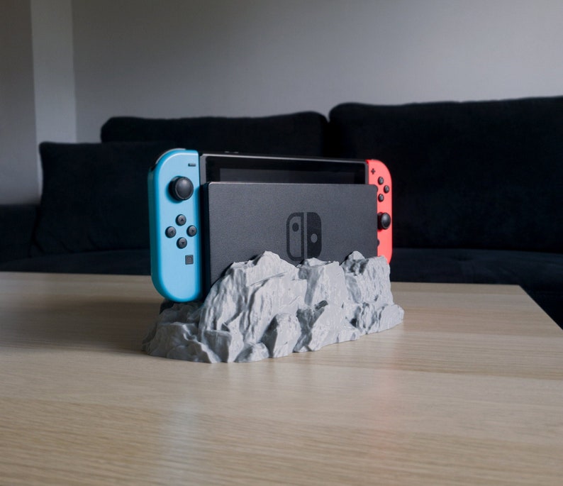 May include: A 3D printed grey rock-shaped stand for a Nintendo Switch console and two controllers. The console is black and the controllers are blue and red.