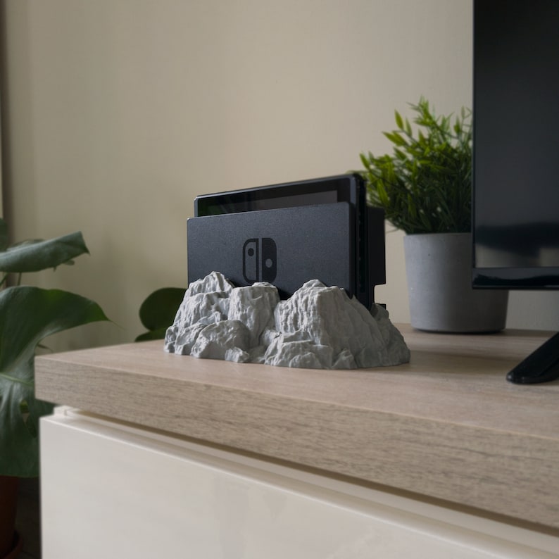 May include: A grey, rocky mountain-shaped stand with a black Nintendo Switch console resting on top. The stand is on a light wood surface.