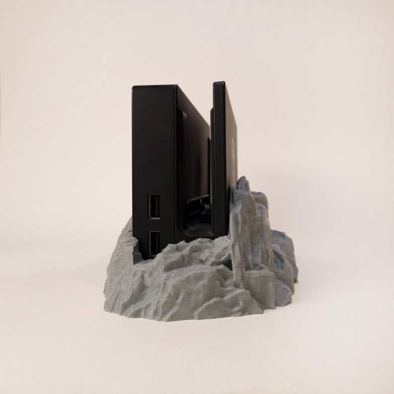 May include: A black Nintendo Switch console and dock stand on a grey, rocky, 3D printed base.