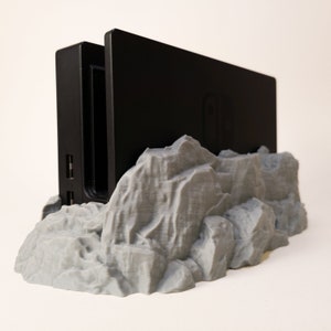May include: A black Nintendo Switch dock sits on a grey 3D printed mountain-shaped stand. The stand has a rough, textured surface.