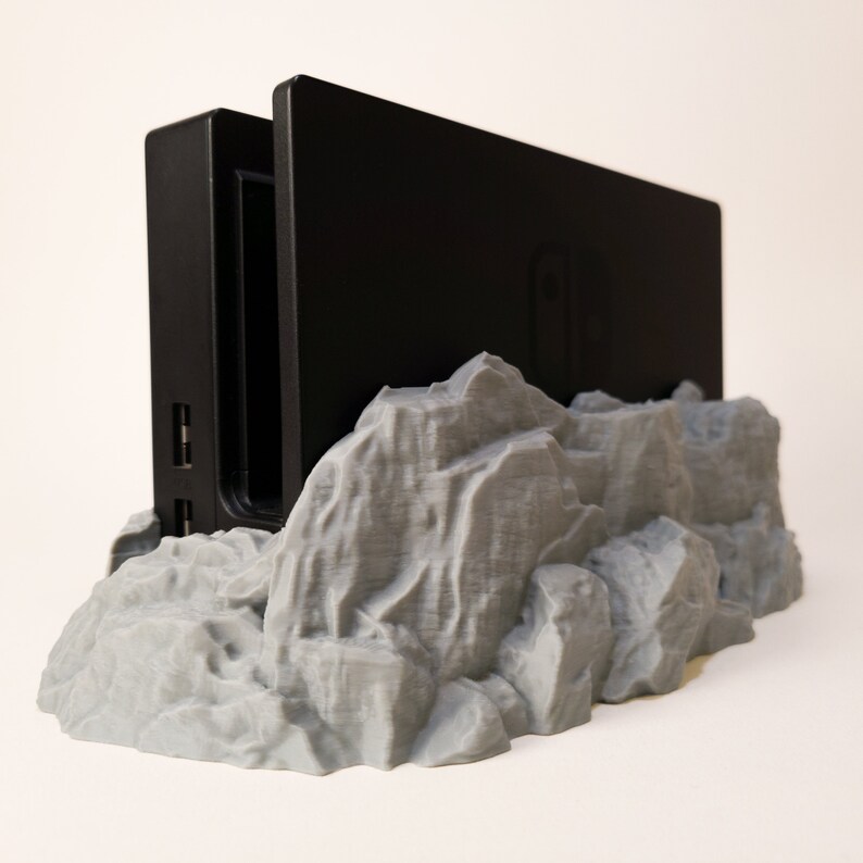May include: A black Nintendo Switch dock sits on a grey 3D printed mountain-shaped stand. The stand has a rough, textured surface.