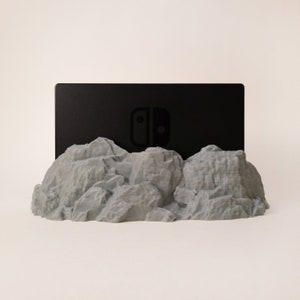 May include: A grey, rocky mountain-shaped stand with a black Nintendo Switch games console resting on top. The console has the Nintendo Switch logo on the front.