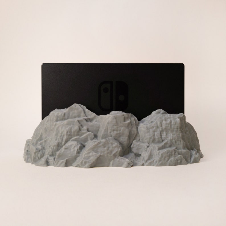 May include: A grey, rocky mountain-shaped stand with a black Nintendo Switch games console resting on top. The console has the Nintendo Switch logo on the front.