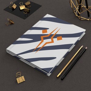 May include: A hardcover journal with a white cover featuring navy blue zebra stripes and an orange geometric design. Gold and black pencils and gold binder clips are arranged around the journal on a dark gray surface.