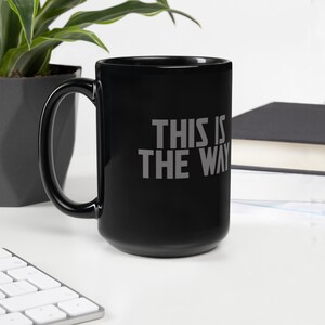 May include: A black ceramic mug with a large handle featuring the gray text THIS IS THE WAY printed on the front. The mug has a smooth, glossy finish and a cylindrical shape, providing a simple and functional design for holding hot or cold beverages.