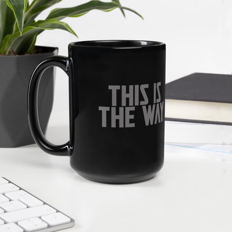 May include: A black ceramic mug with a large handle featuring the gray text THIS IS THE WAY printed on the front. The mug has a smooth, glossy finish and a cylindrical shape, providing a simple and functional design for holding hot or cold beverages.