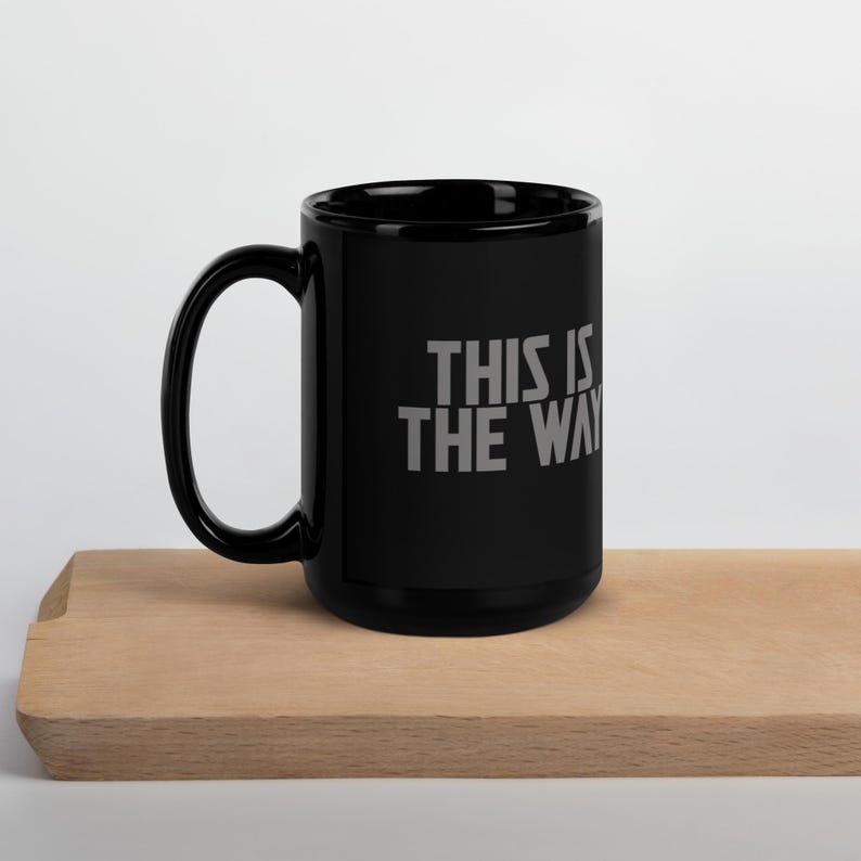 May include: A black ceramic mug with a handle on the left side. The mug features the text "THIS IS THE WAY" printed in a bold, grey, sans-serif font on the front surface. The design is centered on the exterior wall of the cylindrical vessel.