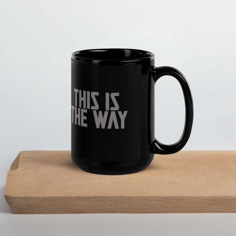 May include: A black ceramic mug featuring the text THIS IS THE WAY printed in a light gray, sans-serif font on the side. The mug has a cylindrical body with a curved handle on the right side and a smooth, glossy finish throughout the exterior surface.