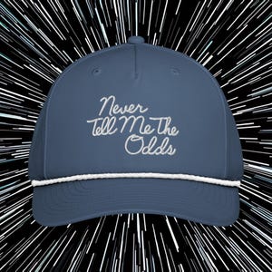 May include: A blue baseball cap with the embroidered text "Never Tell Me The Odds" in white script. The cap features a white rope detail along the brim and a curved visor. The background is a black and white starburst design.