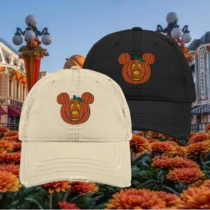 Puede incluir: Dos gorras de béisbol, una crema y otra negra, cada una bordada con una cara de calabaza sonriente con orejas de Mickey Mouse. Las gorras están desgastadas, con un aspecto usado. El fondo presenta follaje otoñal y un entorno de Main Street.
