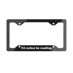 May include: A rectangular black license plate frame with a white speckled pattern throughout. The bottom section features the white text "I'd rather be reading." The frame includes four circular mounting holes positioned at the corners.