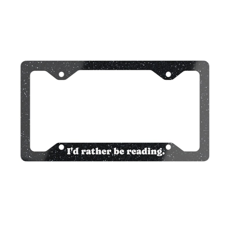 May include: A rectangular black license plate frame with a white speckled pattern throughout. The bottom section features the white text "I'd rather be reading." The frame includes four circular mounting holes positioned at the corners.