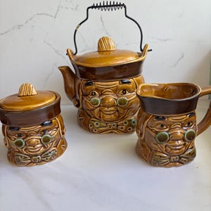 May include: A vintage ceramic tea set featuring a teapot with a black handle, a sugar bowl, and a creamer. The set is brown and tan with a smiling face design, and green-tinted glasses. The teapot has a lid and spout.