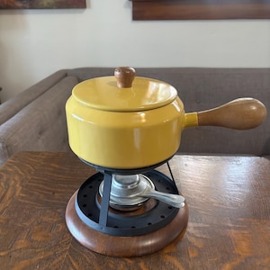 May include: A yellow fondue pot with a wooden handle and lid knob. The pot sits on a black metal stand with a wooden base. A small silver burner is visible. The background includes a painting and a window.