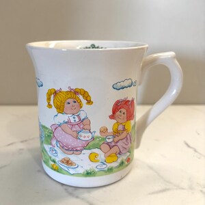 May include: White ceramic mug featuring a colorful illustration of two dolls having a tea party. The dolls have blonde and red hair, wearing dresses. The mug has a curved handle and a slightly flared rim. The design includes clouds and a picnic scene.