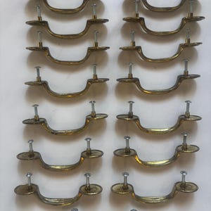 May include: A collection of sixteen brass-toned drawer pulls. Each pull has a curved handle design with two mounting points and a screw. The pulls are arranged in four rows of four, showcasing their symmetrical design and vintage aesthetic.